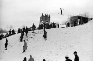 Black and white historical photo of a ski hill in winter, a small crowd of onlookers, and a skiermid-jump, high in the air.