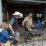 Two men, one in a cowboy hat, at a metal forge with several children looking on, in the bay of a garage.